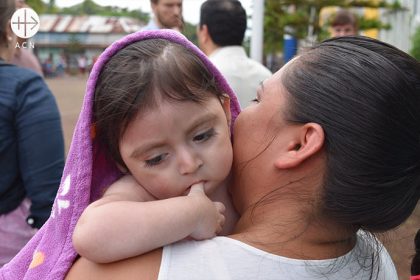 A mother carries her daughter on her arm - wrapped in a blanket. The daughter sucks her thumb.